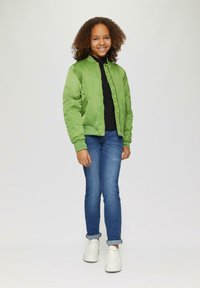 Veste bomber verte avec une texture lisse, pull noir en dessous, jean bleu et baskets blanches. Le design comprend une fermeture éclair à l'avant et des poches.