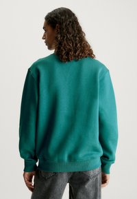 Felpa turchese con colletto a girocollo, polsini e vita a costine; presenta una vestibilità morbida e una texture liscia. I jeans grigi completano l'outfit.