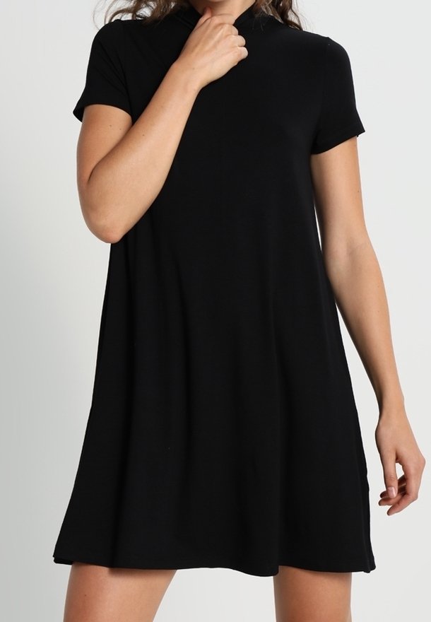 Femme portant une robe noire à manches courtes, longueur au genou, avec une silhouette en ligne A sur un fond clair uni.
