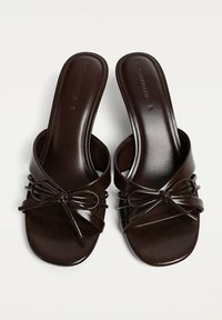Smeđe lažne kožne sandale na slip-on s širokom trakom, ukrasnim mašnom i ravnim đonom. Glatka tekstura s sjajnom završnom obradom.