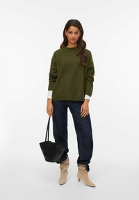 Pull en tricot vert à manches longues, associé à un jean bleu marine et des bottines beiges. Sac à main noir avec une texture douce.