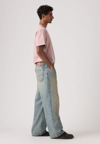 Svetloružové tričko s krátkym rukávom v kombinácii so širokými džínsami vybledlej modrej farby. Nositeľ má ruky vo vreckách a stojí pred jednoduchým pozadím.