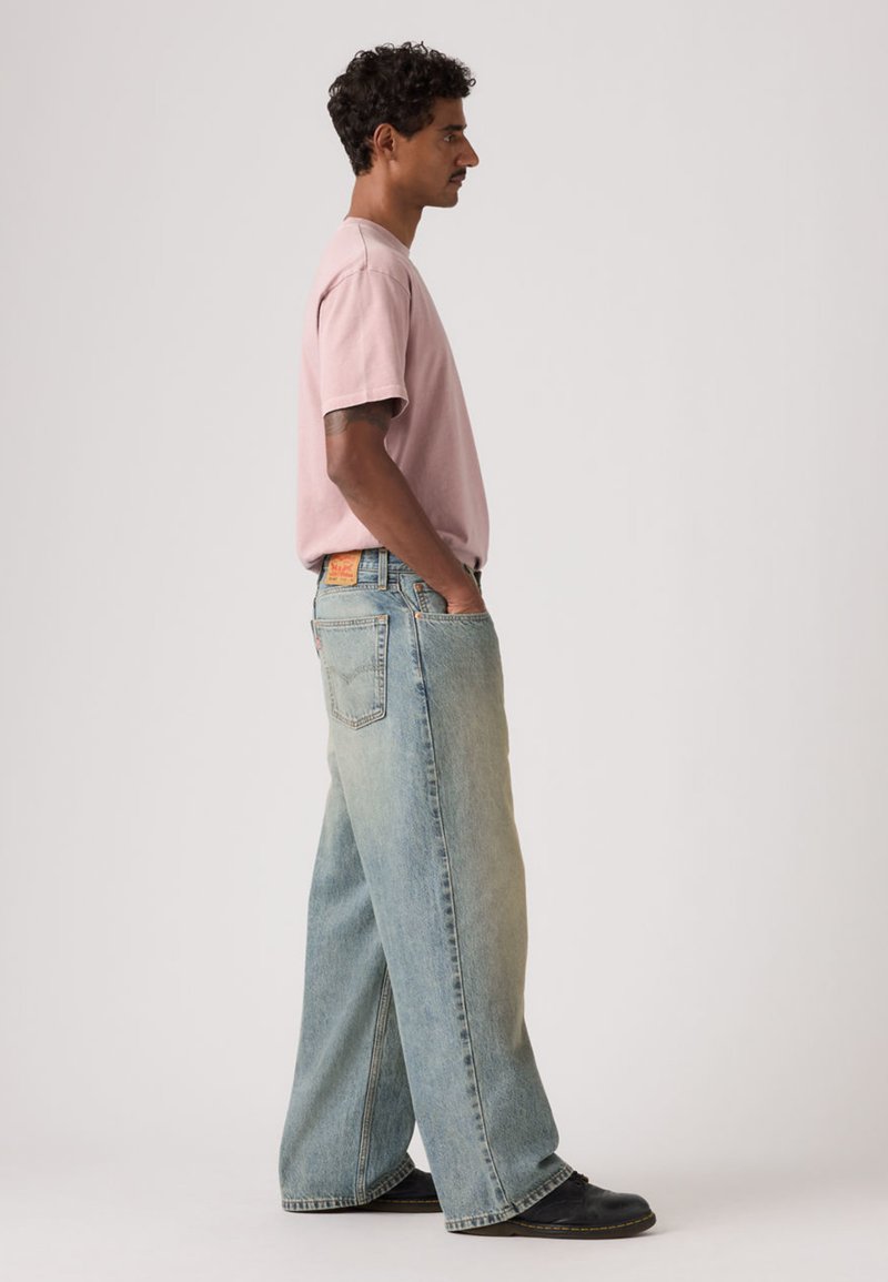 Lyserød t-shirt med korte ærmer kombineret med vidde, falmede blå cowboybukser. Personen har hænderne i lommerne og står foran en ensfarvet baggrund.