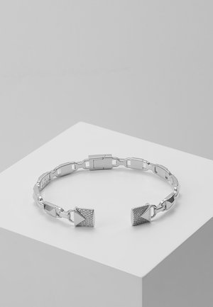 Offenes Silberarmband mit geometrischen quadratischen und dreieckigen Enddetails, präsentiert auf einem weißen Podest vor einem hellgrauen Hintergrund.