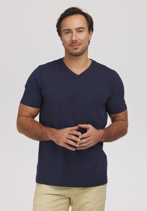 Mężczyzna z krótkimi ciemnymi włosami, ubrany w granatowy T-shirt z dekoltem w serek i beżowe spodnie, stoi ze delikatnie splecionymi palcami na jasnym tle.