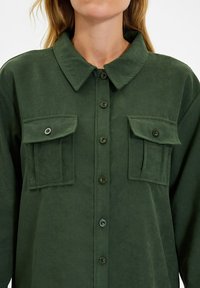 Camicetta verde scuro con bottoni realizzata in tessuto morbido, dotata di colletto, due tasche frontali con pattina e dettagli in bottone neri.