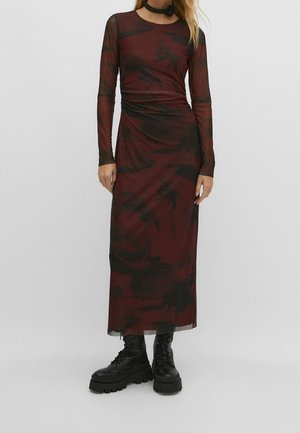 Kobieta w bordowej sukience z długim rękawem sięgającej do kostek, z czarnymi kwiatowymi wzorami, czarnych butach wojskowych i czarnym naszyjniku typu choker.