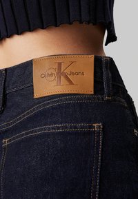 Vaqueros de mezclilla de color azul oscuro, con una etiqueta de cuero marrón claro grabada con "Calvin Klein Jeans" y costuras amarillas en contraste.