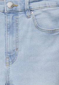 Jean en denim bleu clair en gros plan montrant la poche avant, la boucle de ceinture, le bouton en métal et les détails de couture contrastée.
