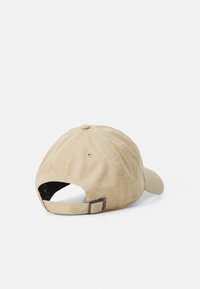 Beige bomullslue med buet skjerm, en knapp på toppen og en justerbar stropp bak med en metallkrok.
