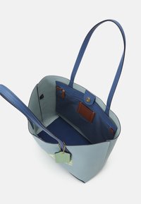 Vaaleansininen olkalaukku, jossa on pitkät kahvat, tummansininen sisätasku vetoketjulla ja vihreä tagi roikkumassa yhdestä kahvasta.