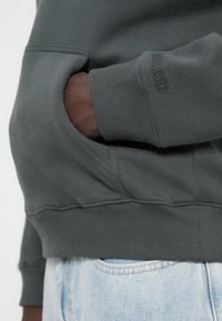 Szara bluza z kangurzą kieszenią, prążkowanymi mankietami i dołem. Materiał ma gładką teksturę z subtelnym logo na rękawie.