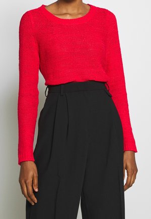 Personne portant un pull en maille rouge vif à manches longues rentré dans un pantalon noir ample taille haute, sur un fond uni.