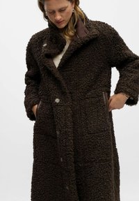 Manteau texturé marron en matériau pelucheux avec un col haut, deux poches avant et un système de fermeture à boutons. Présente des accents en cuir marron.