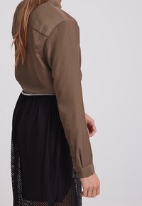 Blusa di seta verde oliva con un yoke posteriore e maniche lunghe, abbinata a una gonna in rete nera con un design aperto a strati.