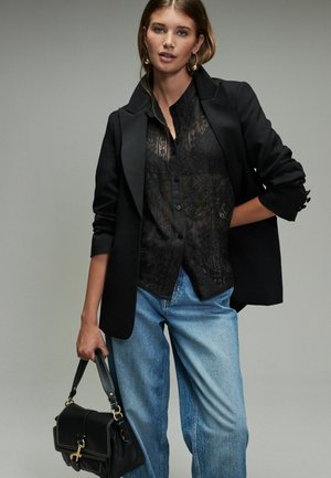 Schwarze Spitzenhemd mit Knopfleiste unter einem taillierten schwarzen Blazer, kombiniert mit hellblauen lockeren Jeans und einer kleinen schwarzen Handtasche mit goldenen Beschlägen.