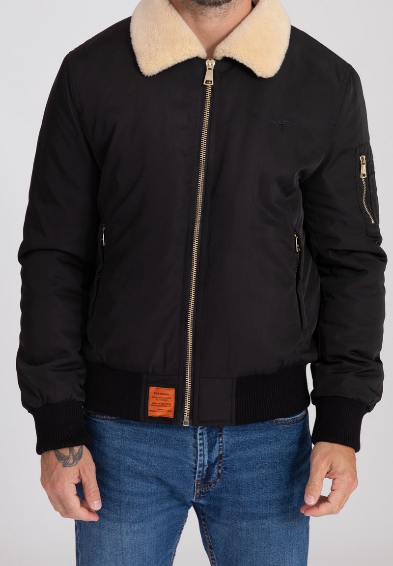 Svart bomberjacka med krämfärgad konstläderkrage, guld dragkedja, ribbade ärmar och en orange lapp vid midjan. Slät yta.