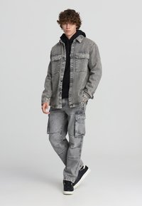 Giacca di denim grigia con grandi tasche, indossata sopra un hoodie nero. Pantaloni cargo grigi con molte tasche e una vestibilità rilassata. Sneakers nere.