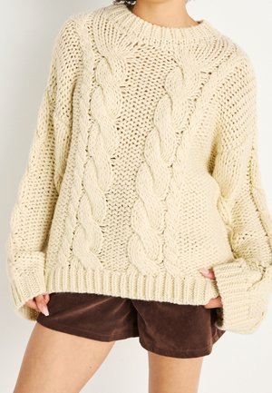 Person iført en chunky cremefarvet kabelstrikket sweater med lange ærmer og brune shorts, hænder delvist stukket ind i sweaterens ærmekanter.