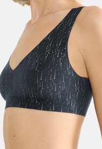 Juodas sportinis liemenėlis su giliu V formos iškirpimu, plačiomis petnešėlėmis ir taškuotais raštais. Pagamintas iš lygios medžiagos, su subtiliu blizgesiu ir minimaliais aksesuarais.