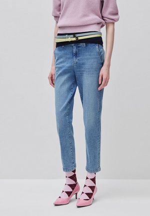 Personne portant un jean bleu clair, un pull rose, deux ceintures, des chaussettes roses et bordeaux à losanges, et des chaussures à talons roses sur un fond uni.