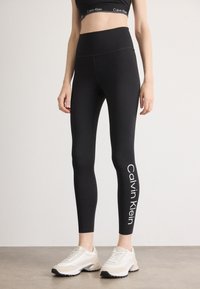 Fekete leggings magas derékkal, a bal lábszáron fehér nyomott logóval. Világos színű sportcipővel párosítva.