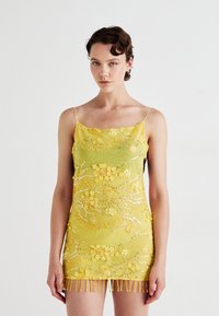 Frau in einem kurzen gelben Kleid mit Spaghettiträgern, floraler Perlenstickerei, Pailletten und Fransenverzierungen am Saum vor einem schlichten Hintergrund.