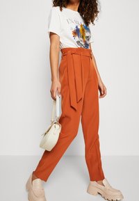 Rostorange högmidjade byxor med ett brett bälte, matchade med en vit grafisk t-shirt och beige plattformsläderskor, hållande en liten vit handväska.