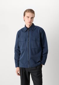 Tumesinine jakk ees oleva lukuga, kahe rinna tasku ja kraega. Valmistatud siledast kangast, millel on lõdvestatud istuvus ja pikad varrukad.