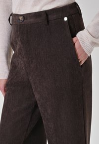 Pantaloni a zigzag marroni con una texture liscia, dotati di chiusura con bottone e tasche laterali. Abbinati a un maglione color crema.