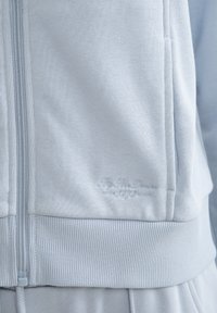 Gaiši zila zip-up jaka ar ribotu piedurknēm un apakšmalu, ar smalku izšūtu logotipu tuvu kabatai. Mīksta, teksturēta auduma.