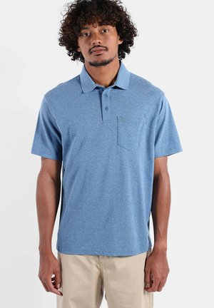 Homme debout face à la caméra, portant un polo à manches courtes bleu avec une poche poitrine et un pantalon beige, sur un fond blanc.