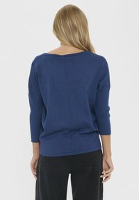 Pulover tricotat bleumarin, cu un decolteu larg, mâneci trei sferturi și o cusătură pe centrul spatelui, purtat cu pantaloni negri.