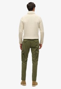 Beige, chunky gebreide coltrui gecombineerd met olijfgroene cargobroek met zijzakken, gedragen met tan kleurige schoenen.
