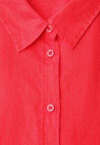 Gros plan sur le col d'une chemise en lin rouge corail avec trois boutons assortis le long de la patte de boutonnage, boutons gravés du texte de la marque.