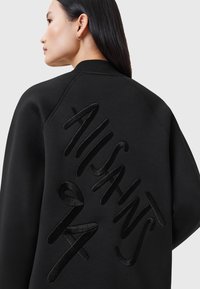 Svart sweatshirt med ribbat krage, med fet, upphöjd text tvärs över ryggen. Slät textur med avslappnad passform.