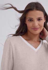 Jasno beżowy sweter z dekoltem w kształcie V z białymi detalami, o miękkiej fakturze, z długimi rękawami i delikatnymi szczegółami. Tło w neutralnym odcieniu.