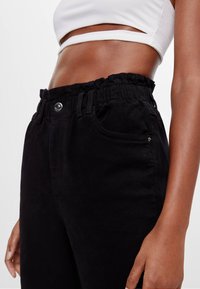 Pantalones de mezclilla negros con una cinturilla elástica, bolsillos frontales y un cierre de botón brillante. La tela tiene una textura suave y un diseño minimalista.