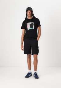 Black graphic t-shirt with a printed design, paired with black cargo shorts. Blue athletic shoes with three white stripes. Black cap.