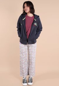 Haut à capuche zippé bleu marine avec des accents verts, t-shirt bordeaux et pantalon large à fleurs. Des baskets noires complètent la tenue.