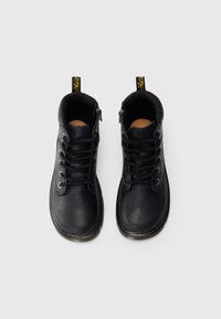 Botas de tornozelo em couro preto com seis ilhós, atacadores pretos, laço no calcanhar amarelo e um detalhe de costura amarela em contraste ao longo da sola.