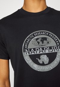 Camiseta de algodón negra con un diseño gráfico circular gris, texto que dice "NAPAPIJRI" y un mapa de la Antártida con un motivo de globo terráqueo.