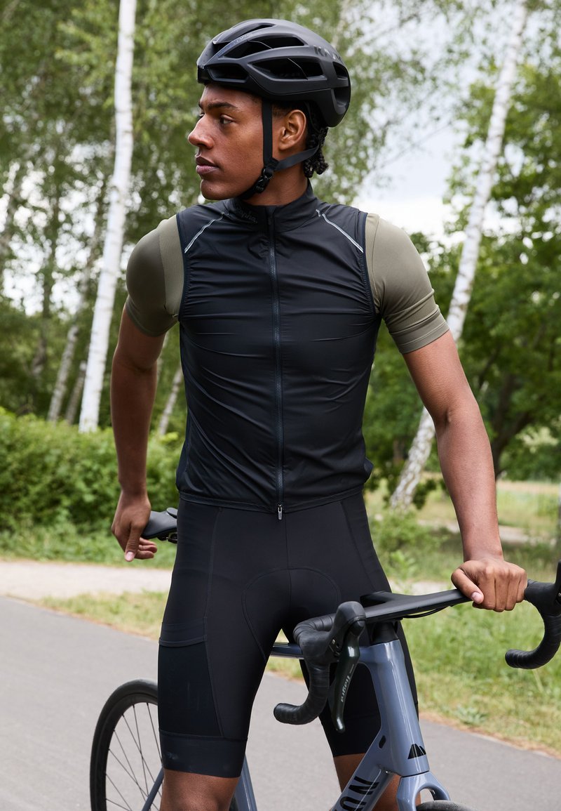 Tenue de cyclisme pour homme avec un maillot sans manches noir et un short sombre. Le casque est noir ; le vélo est gris avec des accents noirs. Fond vert.