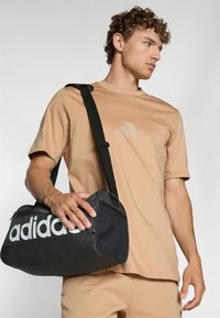 Juodas adidas sportinis krepšys su baltu logotipu, glotnus paviršius ir nuimamas juodas diržas. Modelis vilki šviesiai rudą sportinę marškinių ir šortų komplektą.