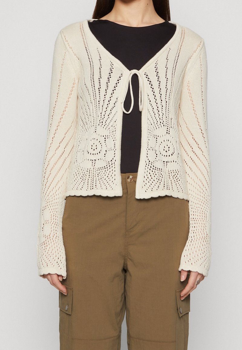 Krämfärgad stickad cardigan med öppet mönster, V-ringning och framknutning. Har blomstermotiv och vågig fåll. Bärs över en mörk topp.