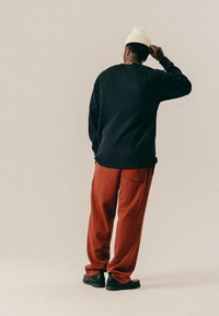 Uomo che indossa un cappello di maglia color crema, un maglione nero e pantaloni color ruggine, in piedi con la schiena rivolta, mentre si sistema il cappello con una mano.