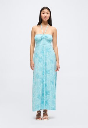 Kvinde iført en lyseblå maxikjole med halterneck og blomstret print samt hvide sandaler med stropper, stående foran en ensfarvet baggrund.