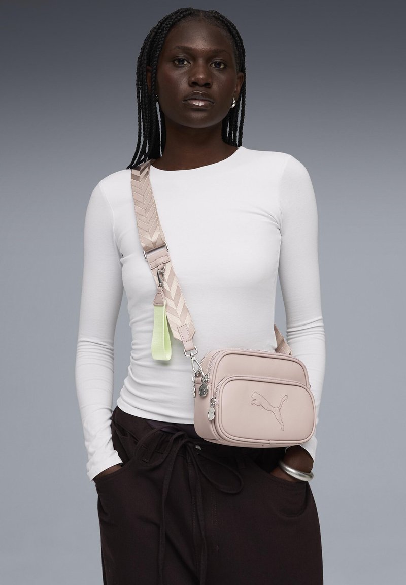 Frau mit geflochtenem Haar, die ein weißes Langarmshirt, schwarze Hosen und eine hellrosa Umhängetasche mit breitem Riemen und Logo auf der Vorderseite trägt.