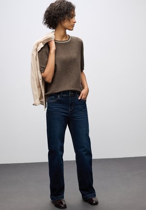 Maglione corto a maniche corte marrone con colletto bianco, abbinato a jeans dritti blu scuro e stivaletti marroni alla caviglia. Giacca leggera gettata su una spalla.