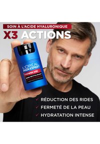 Conteneur cylindrique bleu avec un capuchon de pompe noir, étiqueté "L'Oréal Men Expert Power Age", faisant la promotion des bienfaits du soin de la peau à l'acide hyaluronique.